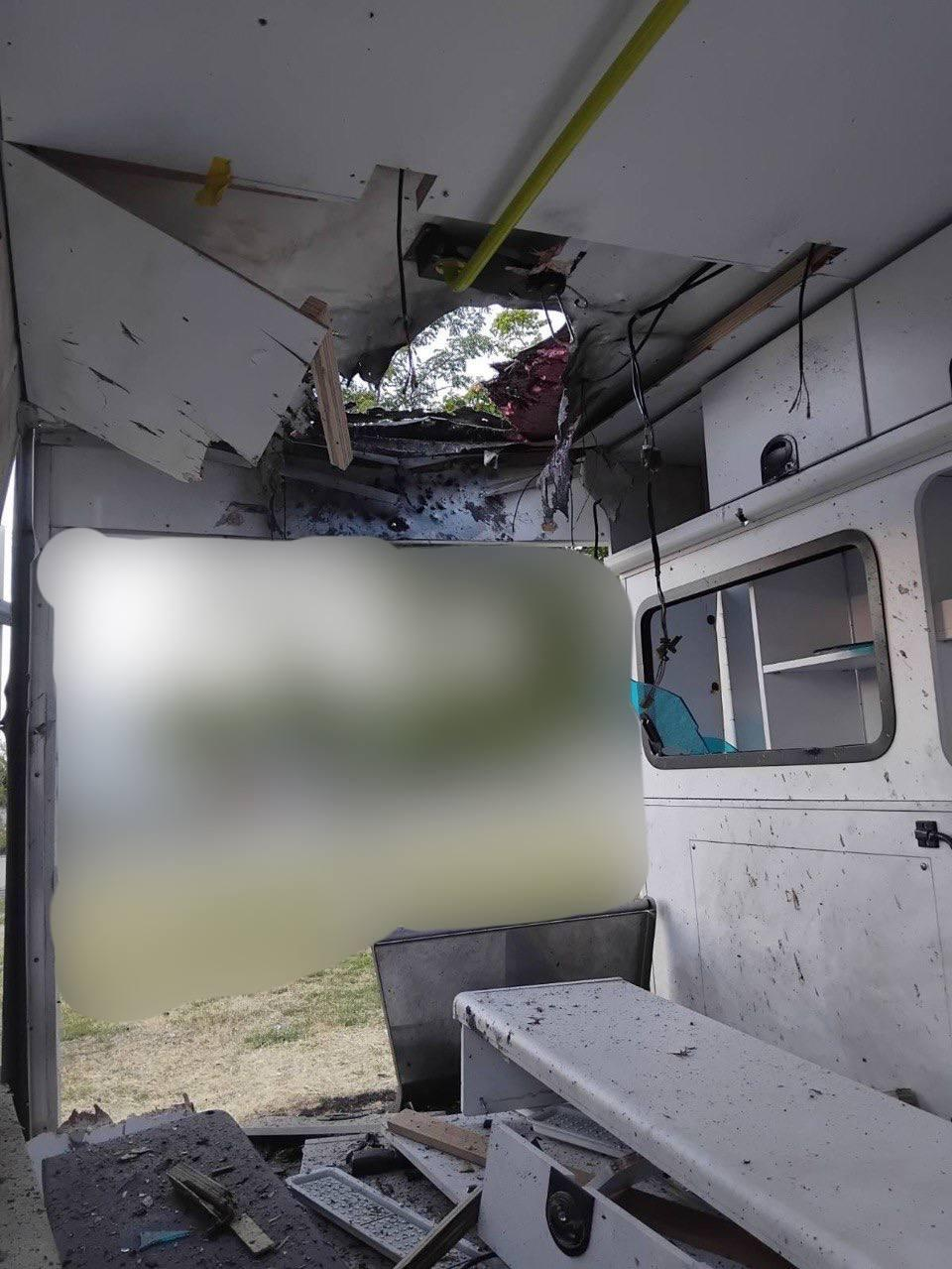 Внутрішній вигляд пошкодженого автомобіля швидкої допомоги із наскрізною вирвою та зруйнованою обшивкою на стелі. Навколо видно розкидані уламки, посічені стіни та вибите скло у внутрішньому віконці медичного відсіку, а ліва частина кадру закрита великою розмитою плямою.