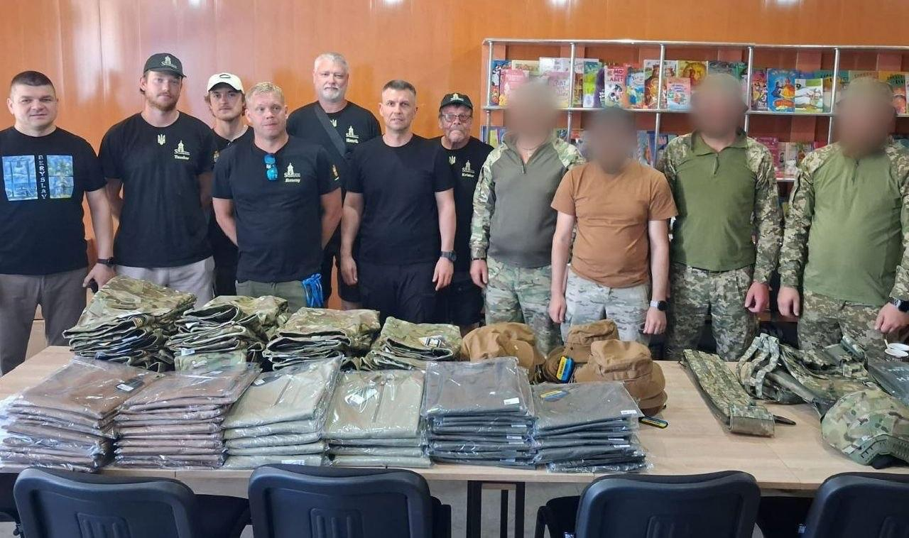 рупа чоловіків стоїть у приміщенні за довгим столом, на якому розкладено велику кількість військової форми та спорядження в упакуваннях і стосах. Частина чоловіків ліворуч одягнена у цивільний одяг та чорні футболки з символікою, а четверо чоловіків праворуч — у камуфляжну тактичну форму (їхні обличчя розмиті з метою конфіденційності). На задньому плані видно книжкові полиці.