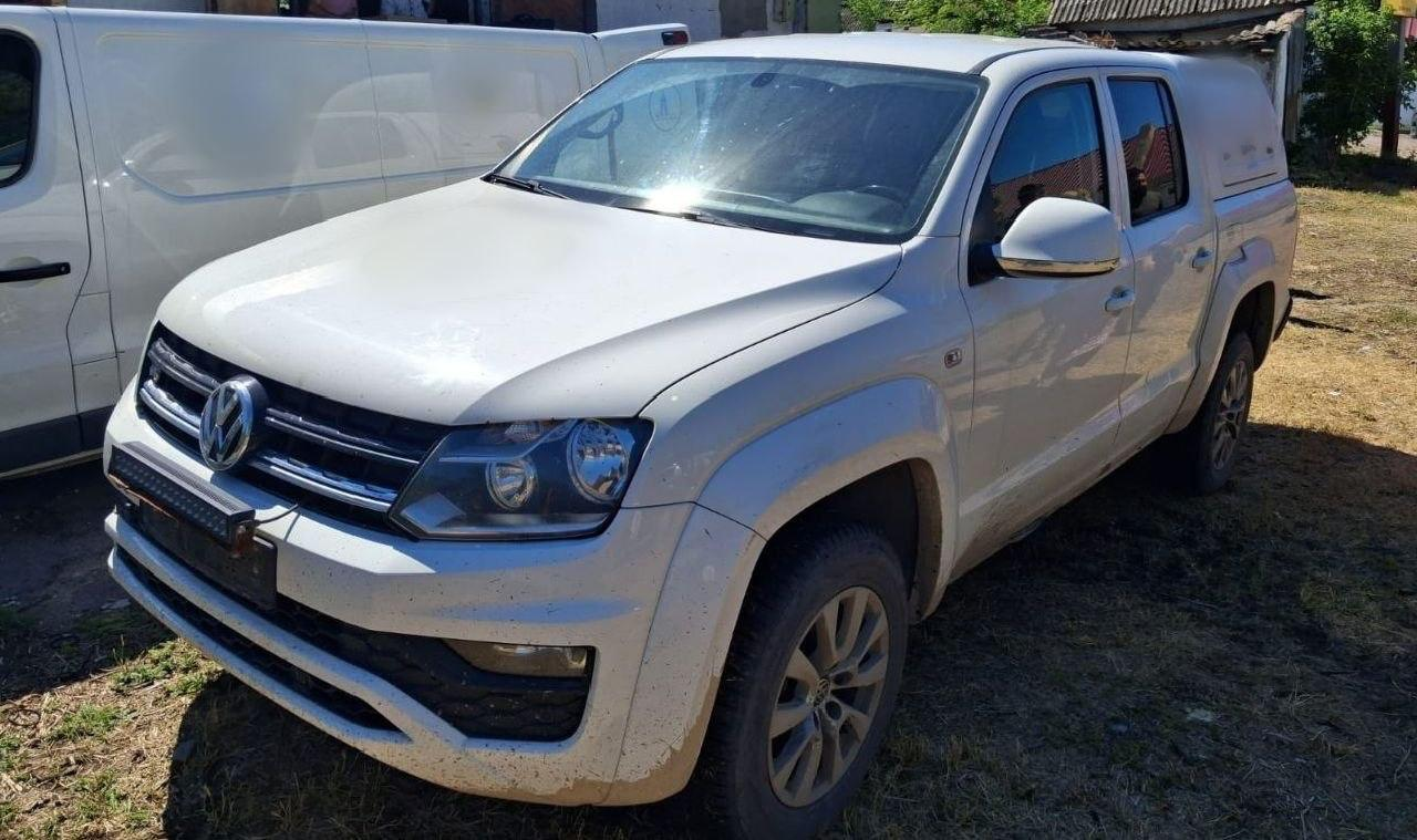 Білий рамний пікап Volkswagen Amarok із кунгом над багажним відділенням стоїть на відкритій місцевості в сонячний день. На передньому бампері автомобіля встановлено додаткову світлодіодну балку (LED-бар), а поруч частково видно інший білий фургон.