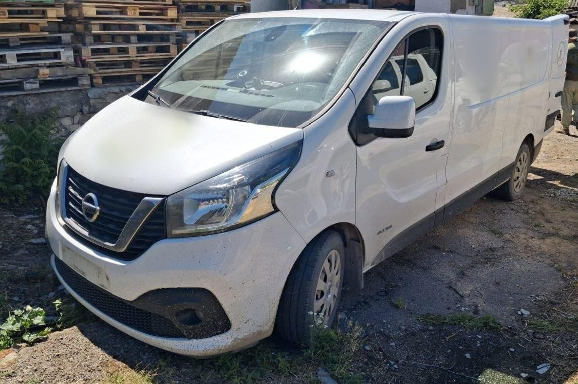 Білий вантажний фургон Nissan Primastar (або схожа модель NV300) стоїть на відкритому майданчику в сонячний день. Автомобіль припаркований під кутом до камери на ґрунтово-асфальтовому покритті, а на задньому плані видно складені дерев'яні піддони (палети).