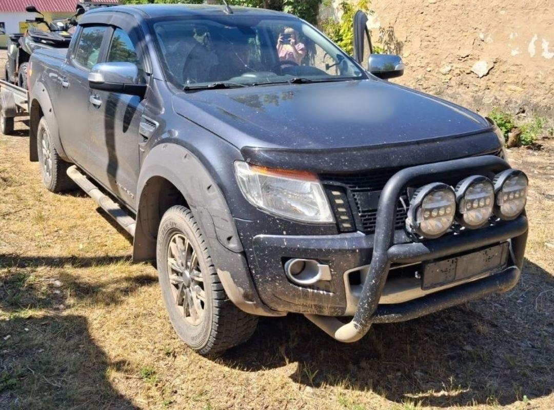 Темно-сірий рамний пікап Ford Ranger із розширеними колісними арками та захисним кенгурятником стоїть на трав'янистому майданчику в сонячний день. На кенгурятнику встановлено три додаткові круглі прожектори, а позаду автомобіля видніється причіп.
