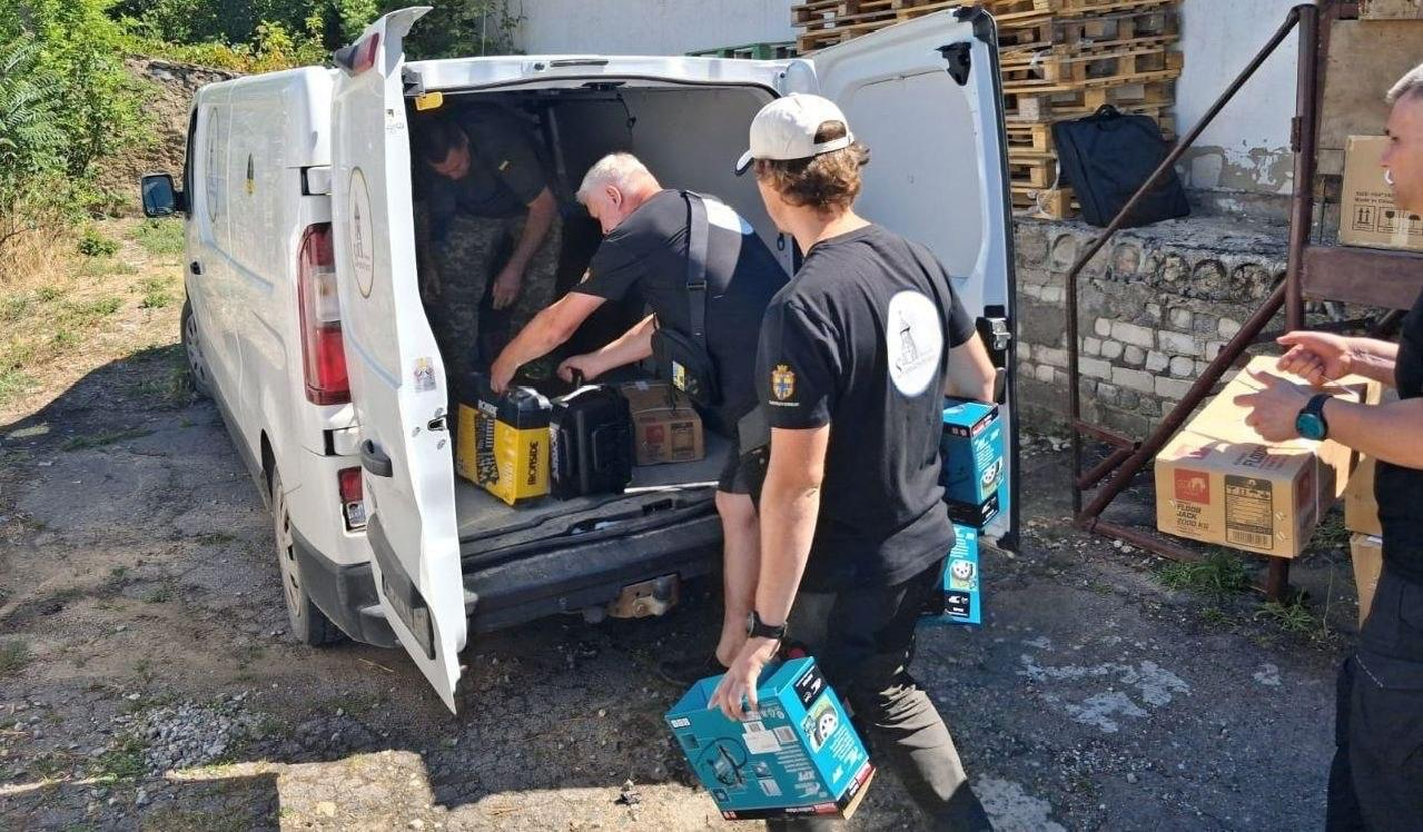 Чоловіки розвантажують або завантажують коробки та обладнання через відкриті задні двері білого вантажного фургона. Один із чоловіків тримає в руках бірюзові коробки з інструментами, а інші всередині та біля кузова переставляють генератор та інші речі. На задньому плані видно дерев'яні піддони та картонні коробки.