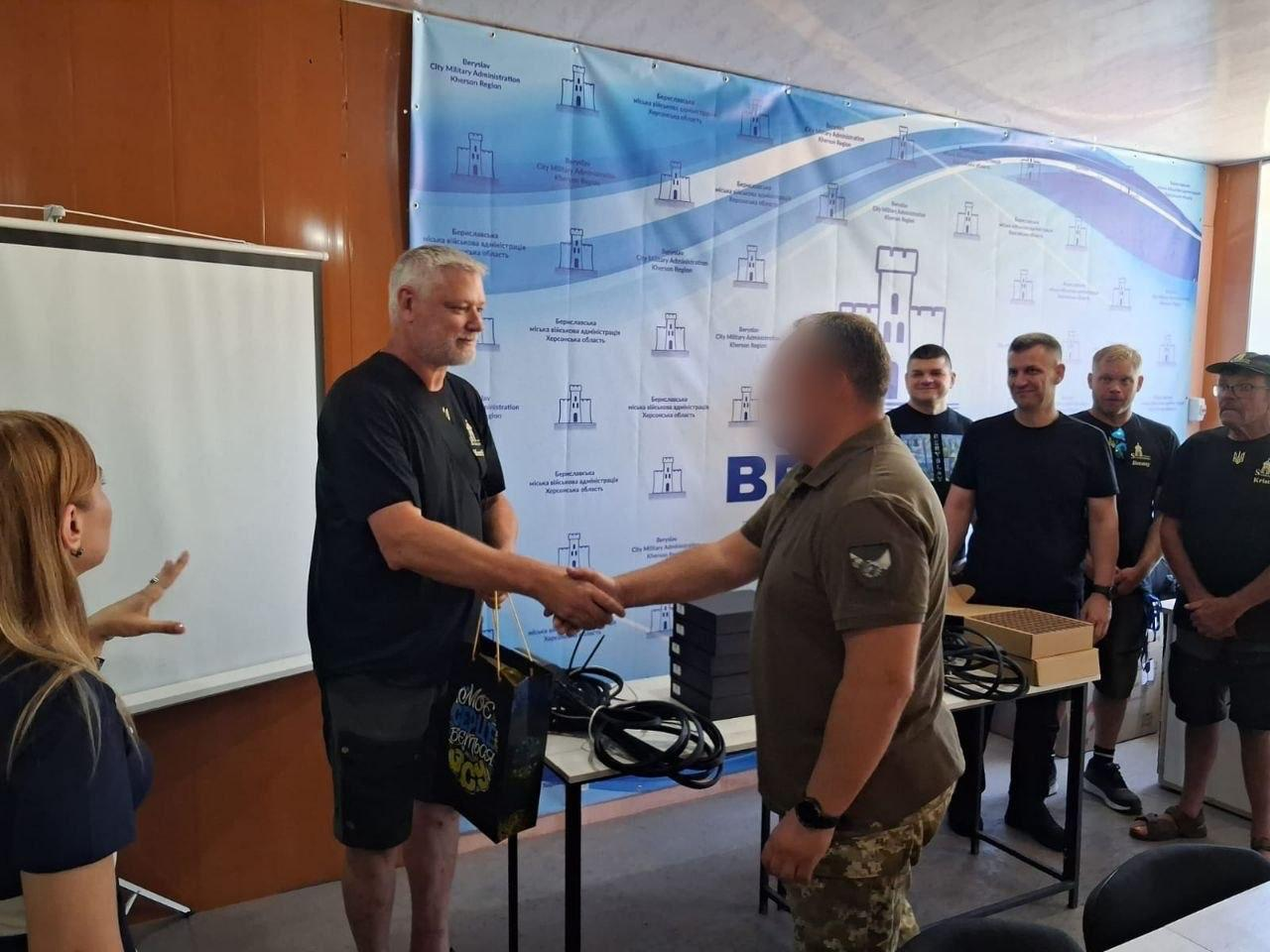 Офіційна зустріч у приміщенні на тлі банера Бериславської МВА. Сивочолий чоловік у чорній футболці тисне руку військовослужбовцю у камуфляжних штанах та футболці кольору хакі (його обличчя розмите з метою конфіденційності) та передає йому подарунковий пакунок. На задньому плані за процесом спостерігають інші чоловіки, а праворуч на столі лежать коробки та кабелі.