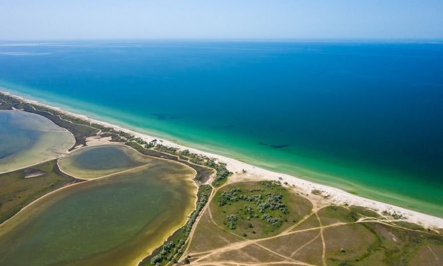 Панорамний знімок піщаної коси з висоти пташиного польоту. Вона розділяє глибоке синє море з бірюзовою водою біля берега та мілководні зеленкуваті лимани. На самій косі видно піщаний пляж, рідкісну зелену рослинність та мережу ґрунтових доріг під чистим блакитним небом.