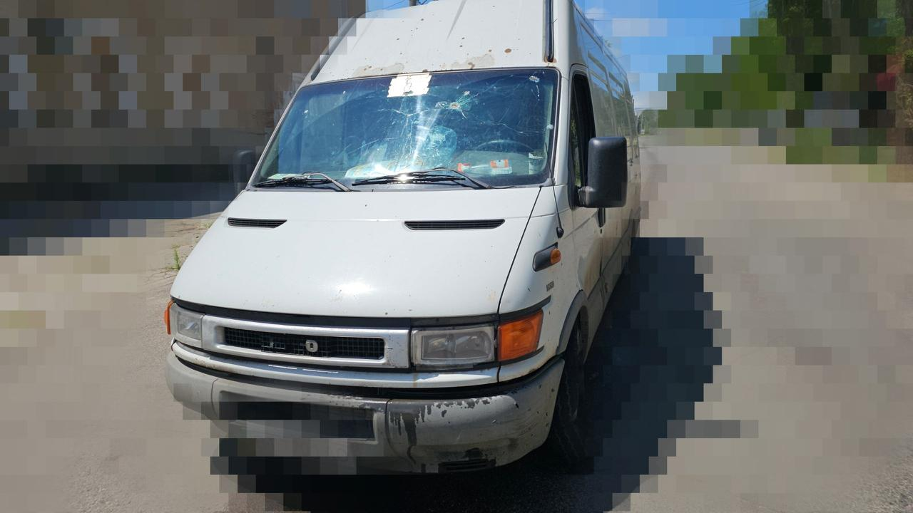 Білий вантажний мікроавтобус (мінівен) стоїть на дорозі в сонячний день. На лобовому склі автомобіля видно значні пошкодження, тріщини та сколи, схожі на сліди від ударів або уламків. Навколо транспортного засобу накладено ефект піксельного розмиття для приховування деталей навколишнього середовища.