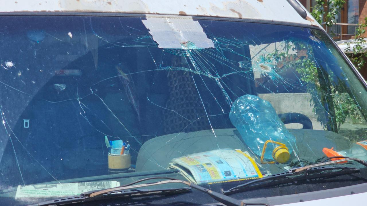 Крупний план пошкодженого лобового скла автомобіля білого кольору. Скло вкрите густою сіткою тріщин, а зверху по центру наклеєно шматок армованого скотчу. Всередині салону на панелі приладів лежать відкритий журнал, порожня велика пластикова пляшка для води, склянка з канцелярією та інші дрібні речі.