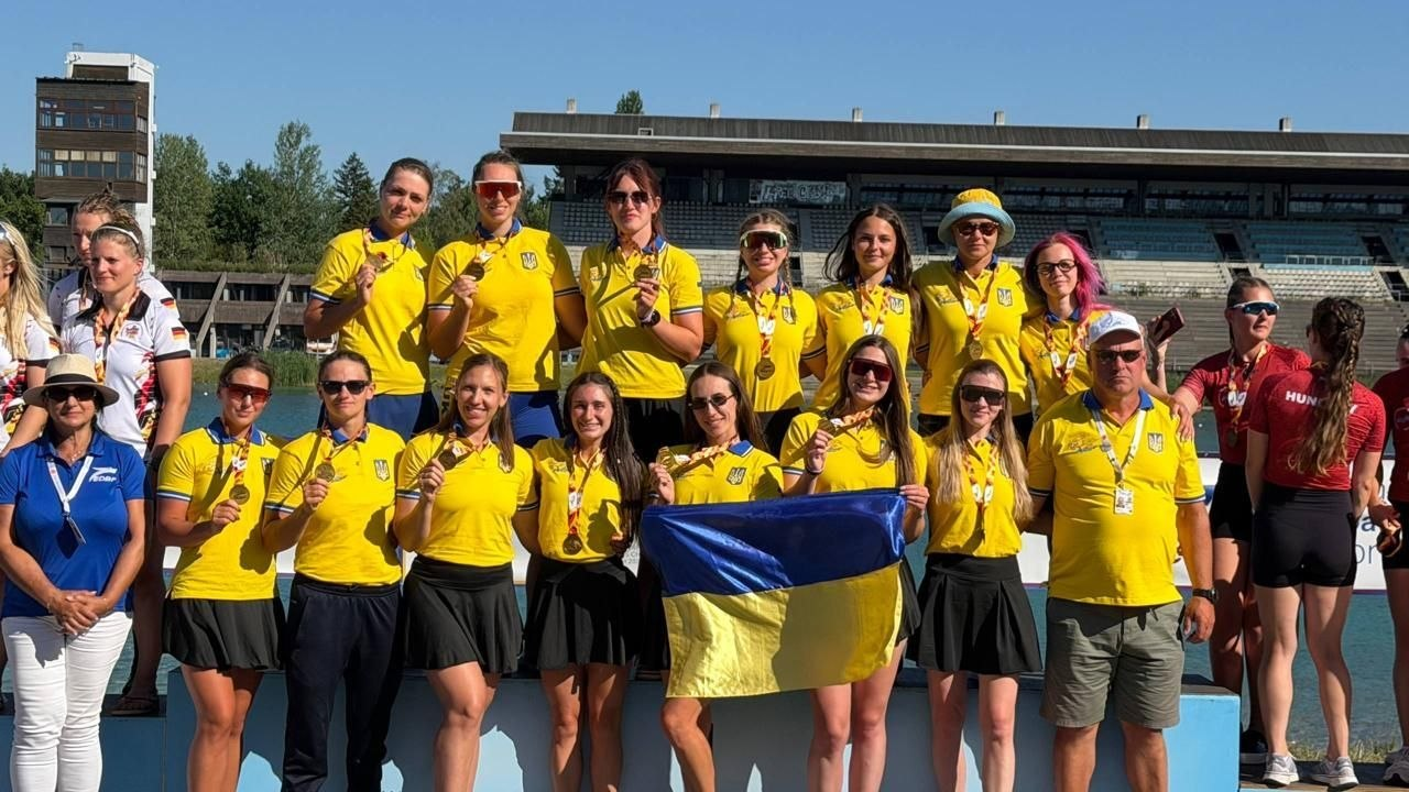 Група спортсменок та тренерів у жовтих футболках збірної України позує з медалями на п'єдесталі пошани під час церемонії нагородження на веслувальному каналі. Попереду вони тримають синьо-жовтий прапор України, а праворуч і ліворуч на п'єдесталі частково видно представниць інших збірних. На задньому плані розташовані трибуни стадіону.