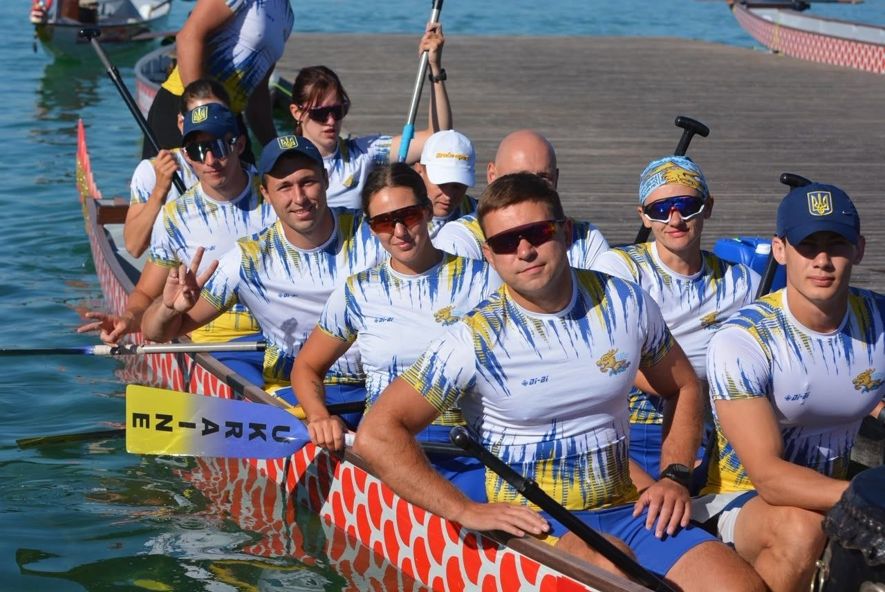 Група спортсменів у біло-синьо-жовтій формі збірної України сидить у великому човні-драконі («драгонботі») біля дерев'яного пірса. Вони тримають весла, на одному з яких є напис Ukraine, та позують для фото. Подія відбувається на відкритій водоймі у сонячний день.