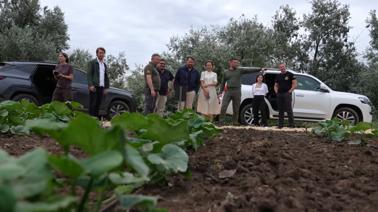 Начальник обласної військової адміністраціїОлександр Прокудін у тактичній формі кольору хакі вказує рукою вбік, спілкуючись із представниками делегації на краю поля. Поруч із присутніми стоїть його перший заступник Дмитро Бутрій, також у тактичній формі, а на передньому плані видно грядки із зеленими рослинами.
