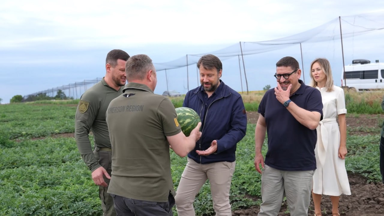 Група людей стоїть на кавуновому полі в похмурий день. Перший заступник начальника ХОВА Дмитро Бутрій у тактичній футболці-поло з написом Kherson Region спиною до камери передає великий кавун чоловікові в синій куртці. Поруч стоять начальник ХОВА Олександр Прокудін в тактичній формі кольору хакі, чоловік в окулярах та чорній футболці-поло, а також жінка у світлій сукні.
