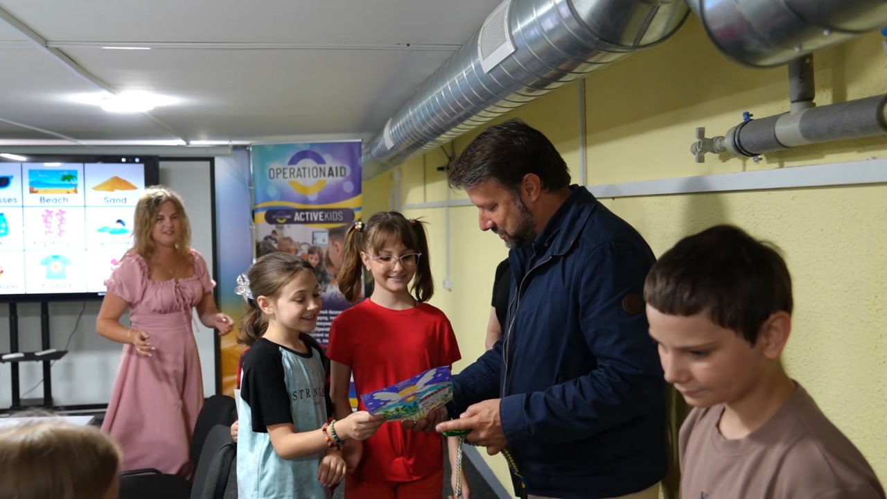 Чоловік у синій куртці спілкується з дітьми у облаштованому приміщенні з вентиляційними трубами під стелею. Дві дівчинки посміхаються та тримають у руках кольоровий дитячий малюнок. На задньому плані видно жінку у рожевій сукні, великий інтерактивний екран із малюнками та банери благодійних організацій.