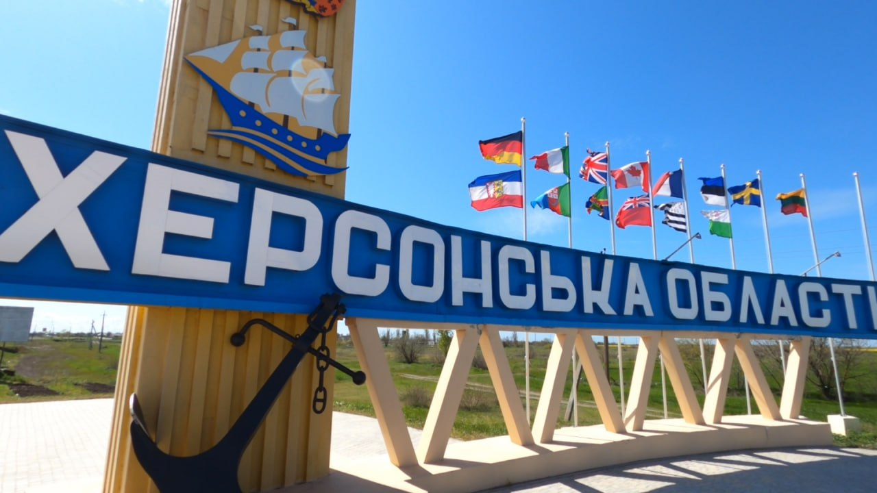 Велика в'їзна стела з написом «ХЕРСОНСЬКА ОБЛАСТЬ» на синьому тлі під яскравим блакитним небом. Конструкція прикрашена зображенням вітрильника та великим чорним якорем біля основи. На задньому плані в ряд височіють флагштоки з прапорами різних іноземних держав, які майорять на вітрі.