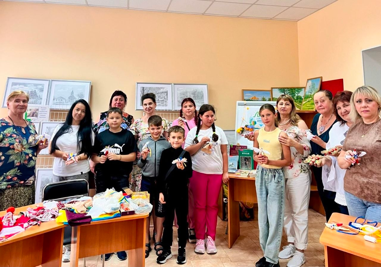 Група дітей та жінок позує для спільного фото в світлій кімнаті під час творчого заходу або майстер-класу. Учасники тримають у руках виготовлені власноруч патріотичні сувеніри, ляльки-мотанки та обереги. На столах попереду розкладені матеріали для творчості й кольорові стрічки, а на задньому плані на стіні розвішані картини та малюнки у рамах.