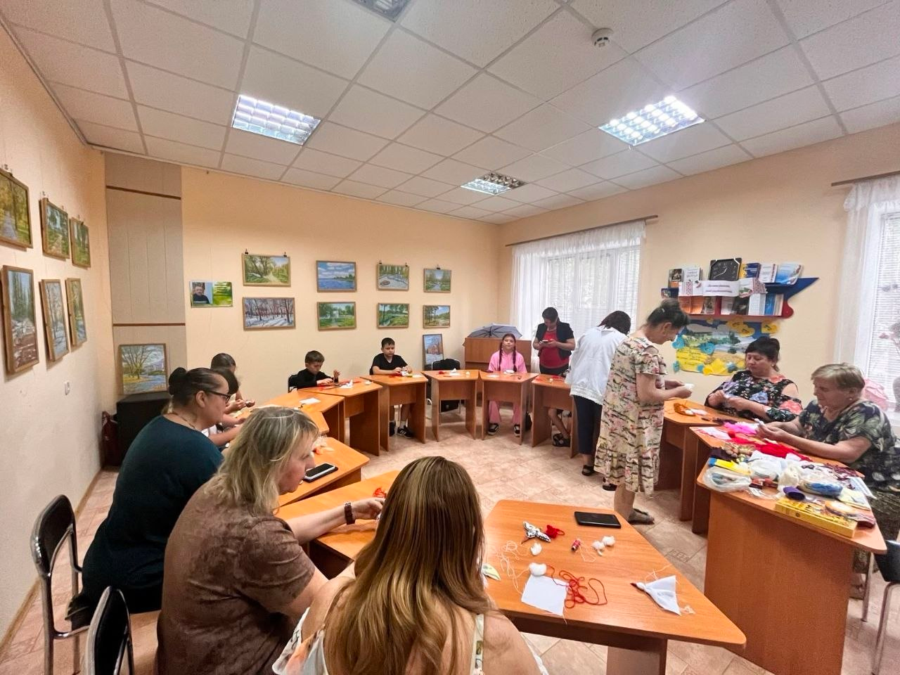 Група дітей та дорослих сидить за напівкруглими столами у просторій кімнаті під час рукодільного майстер-класу. Учасники зосереджено виготовляють вироби з ниток та тканин, а одна з жінок допомагає їм, стоячи у центрі. Стіни приміщення прикрашені численними невеликими картинами у рамах, а на задньому плані праворуч облаштовано книжкову поличку.