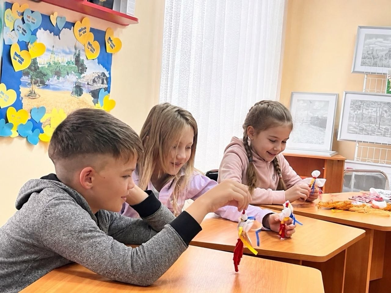 Троє дітей сидять за партами у світлій кімнаті та захоплено граються власноруч зробленими патріотичними ляльками-мотанками з ниток. Хлопчик ліворуч та дві дівчинки посміхаються, розглядаючи свої вироби. На задньому плані видно стіну з дитячим настінним плакатом, прикрашеним паперовими жовто-блакитними сердечками, та картини у рамах.