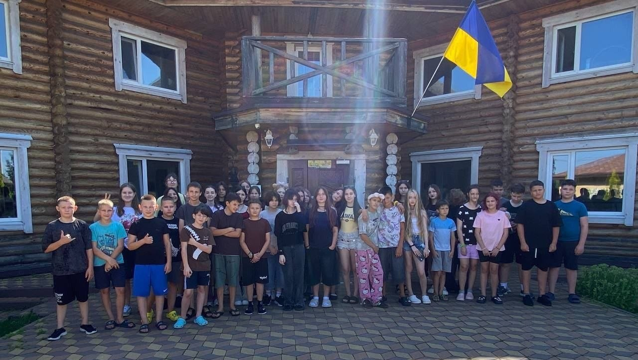 Велика група дітей та підлітків стоїть на подвір'ї перед двоповерховим дерев'яним будинком зі зрубу, над входом якого майорить державний прапор України.