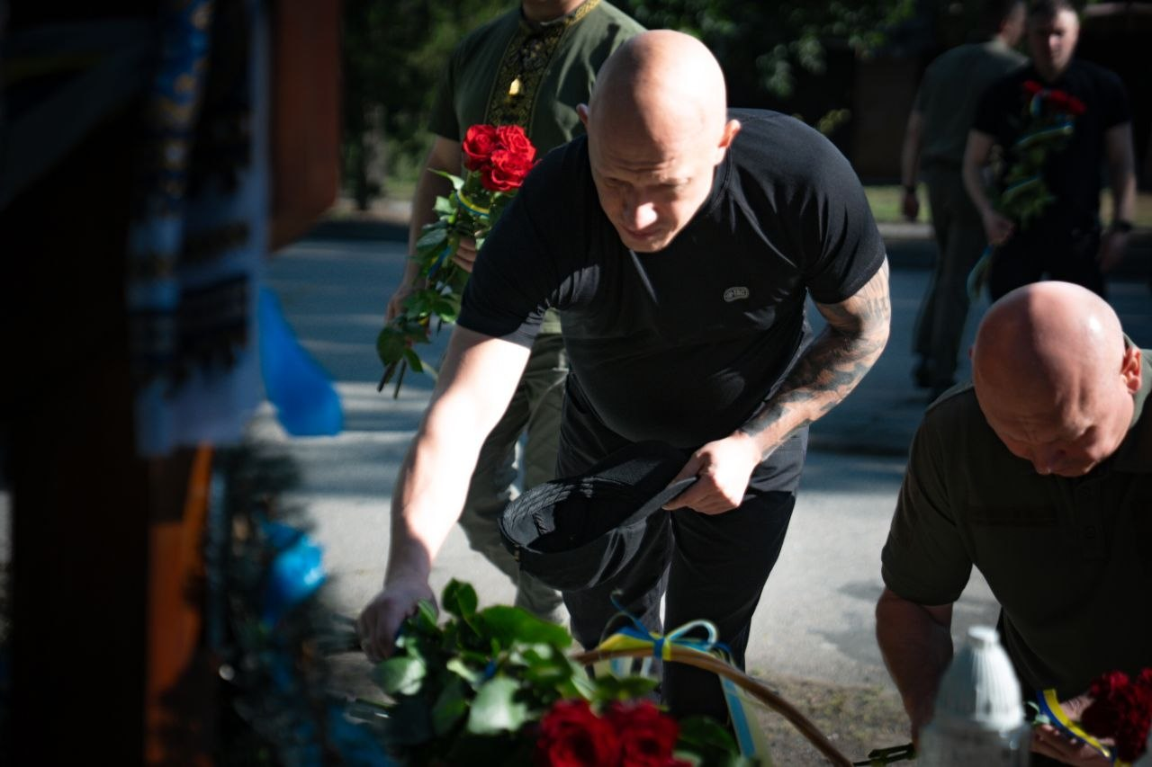 Заступник начальника Херсонської ОВА Євген Ігнатенко у чорній футболці з татуюванням на руці схиляється, щоб покласти квіти до меморіалу пам'яті. У долонях він тримає головний убір. Поруч та на задньому плані зображено керівництво Херсонської ОВА у військовій формі та вишиванках, які також тримають червоні троянди.
