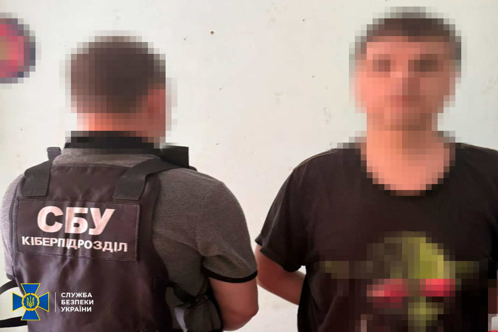 Співробітник СБУ затримує підозрюваного. Ліворуч спиною до камери стоїть правоохоронець у бронежилеті з написом «СБУ КІБЕРПІДРОЗДІЛ». Праворуч обличчям до камери стоїть затриманий чоловік у чорній футболці, його руки заведені за спину. Обличчя обох чоловіків розмиті (пікселізовані).