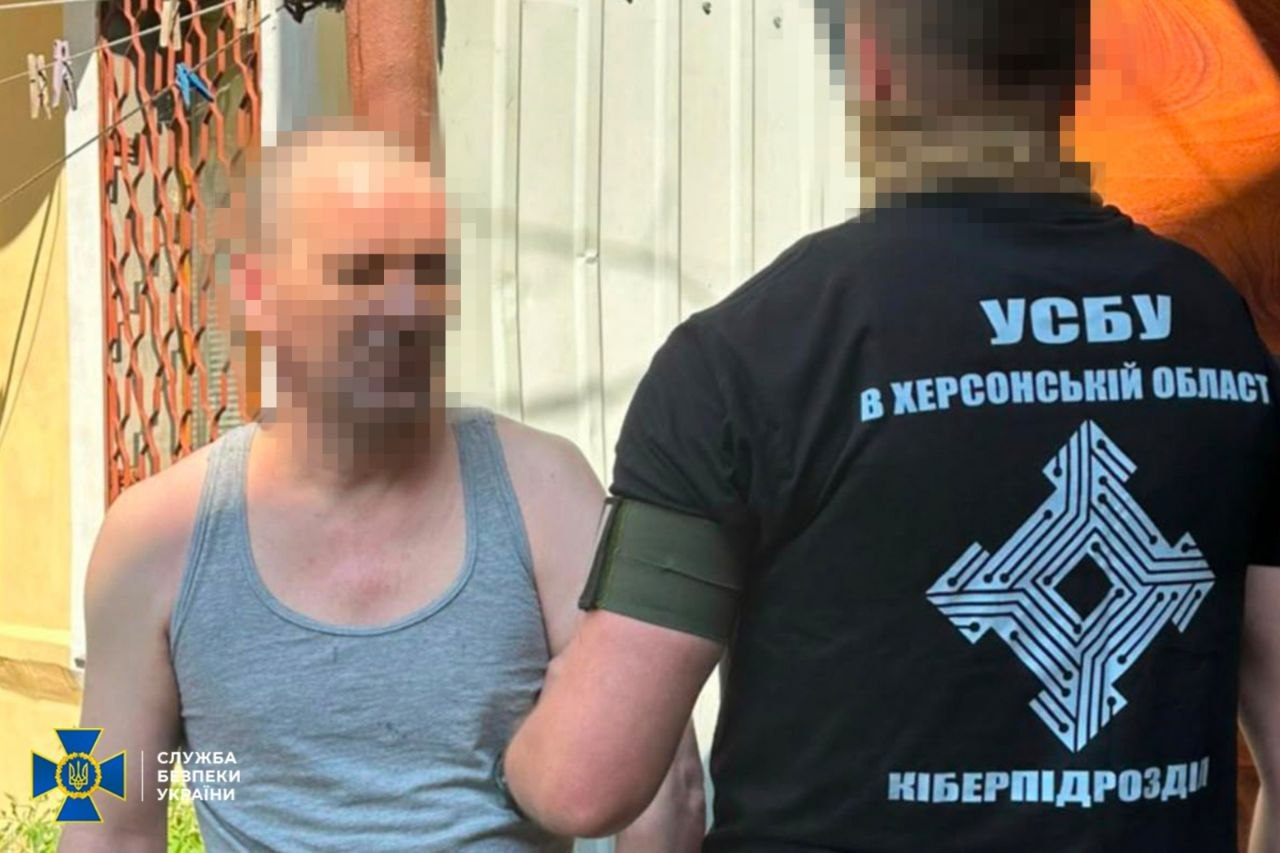 Співробітник СБУ затримує підозрюваного в Херсонській області. Праворуч спиною до камери стоїть правоохоронець у чорній футболці з емблемою та написом «УСБУ в Херсонській області Кіберпідрозділ». Ліворуч обличчям до камери стоїть затриманий чоловік у сірій майці, його руки заведені за спину. Обличчя обох чоловіків пікселізовані.
