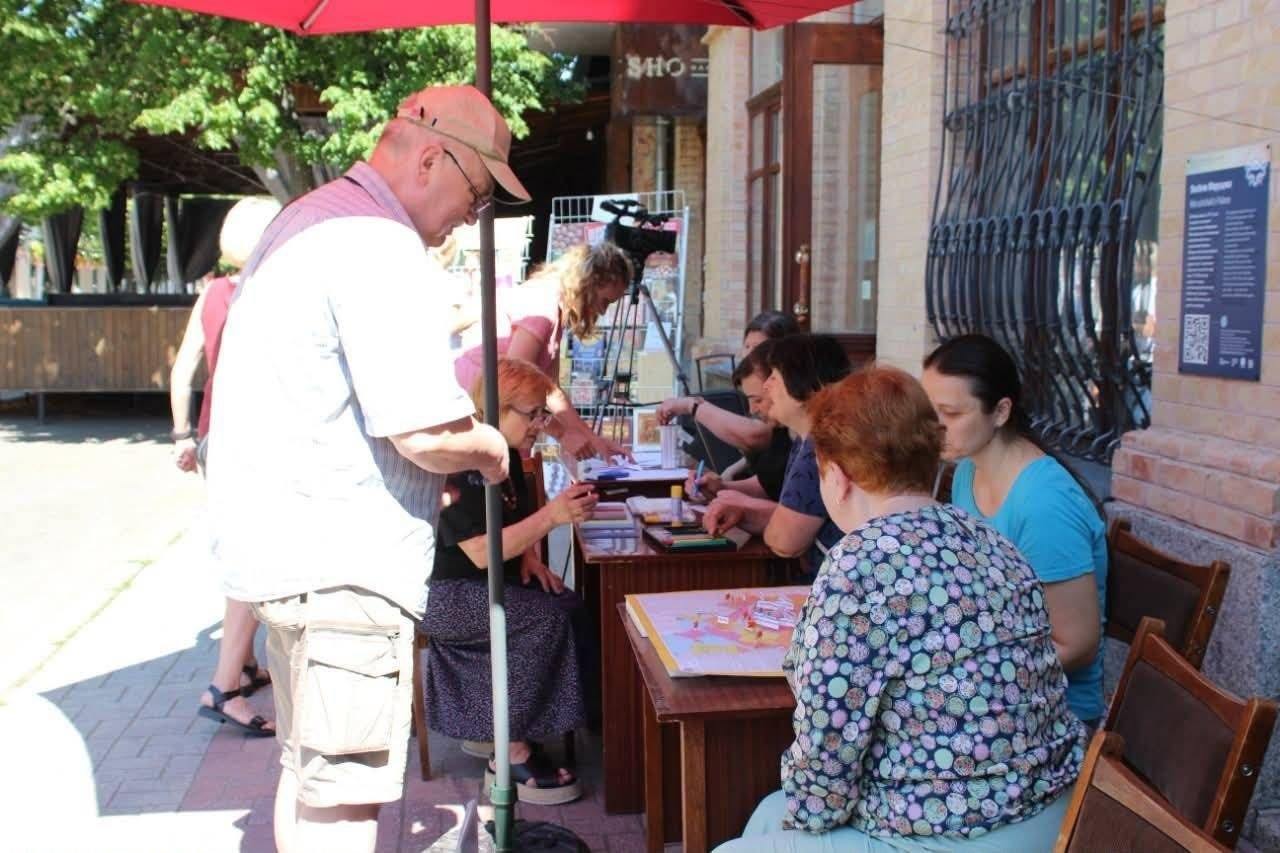 Майстер-клас або громадський захід на вулиці міста. Під великою червоною парасолькою облаштовано столи, за якими жінки заповнюють документи, малюють або працюють із мапами. Ліворуч біля столу стоїть літній чоловік у світлій сорочці, шортах та кепці, який спілкується з організаторами.