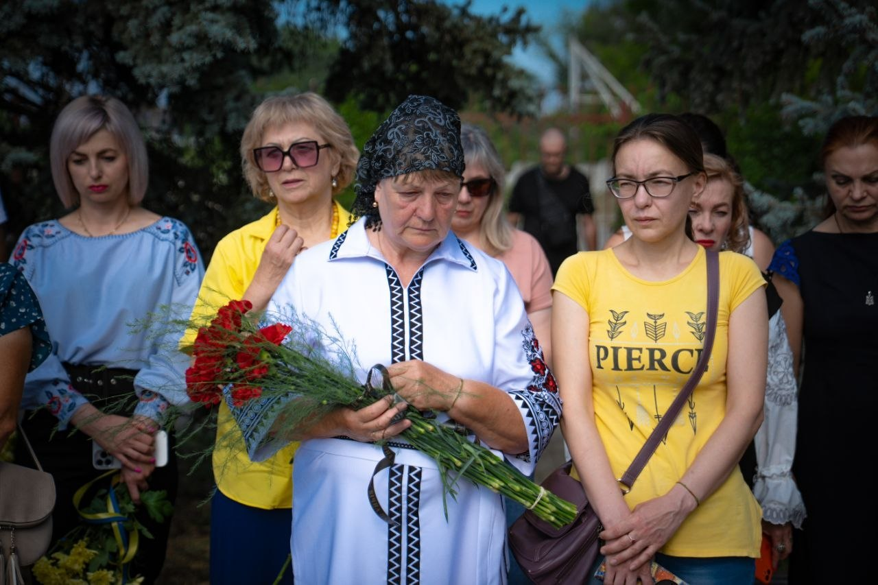 Учасники заходу пам'яті зі скорботою на обличчях. У центрі стоїть літня жінка у білій вишиванці з хусткою на голові, яка тримає букет червоних гвоздик. Справа від неї стоїть дівчина в окулярах та жовтій футболці, а зліва — жінка в сонцезахисних окулярах і жовтій сорочці. На задньому плані присутні інші люди в жалобі.