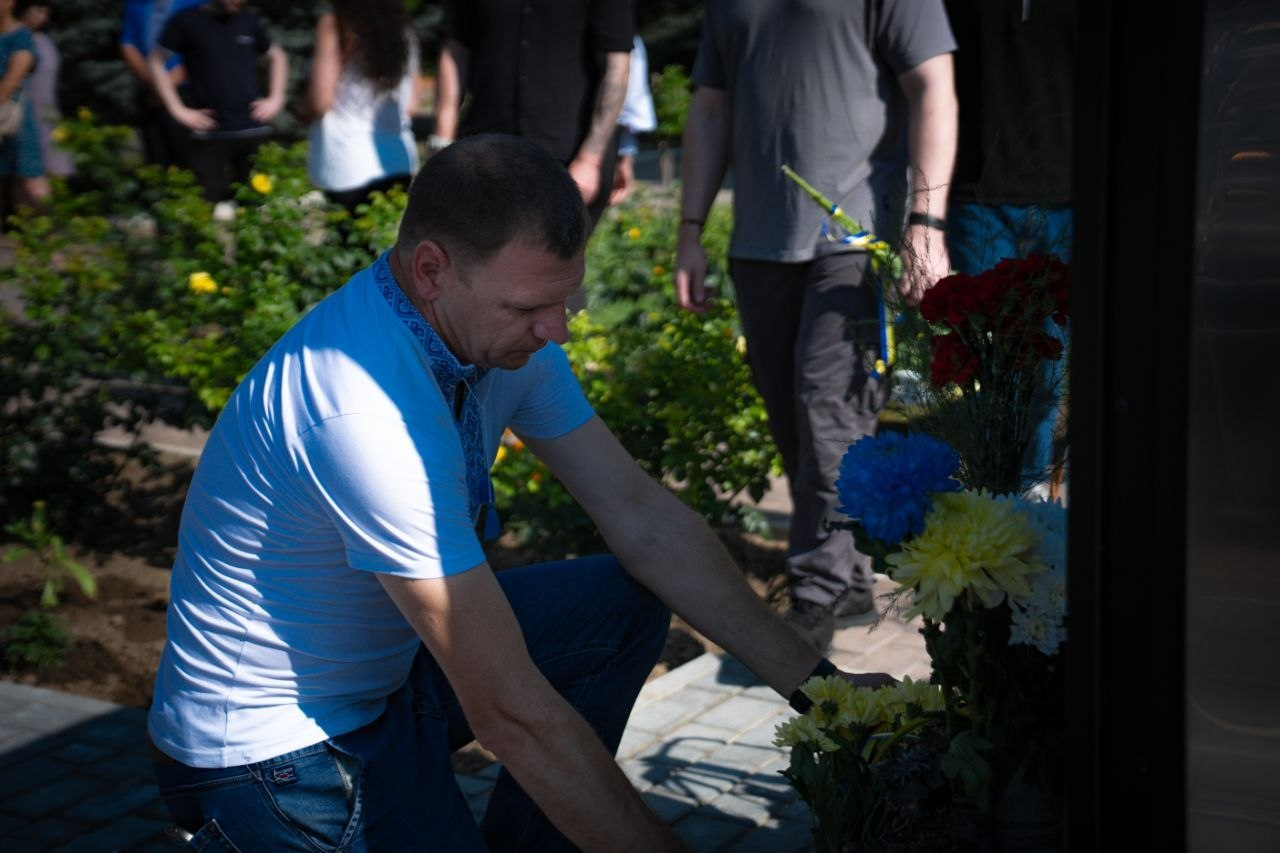 Начальник Генічеської МВА Євгеній Родіонов покладає квіти до меморіалу. Чоловік у світло-синій футболці з вишитим коміром та в джинсах приклонив коліно перед пам'ятним знаком. Він поправляє синьо-жовті хризантеми та червоні троянди, покладені біля підніжжя. На задньому плані видно інших учасників заходу в скорботі.