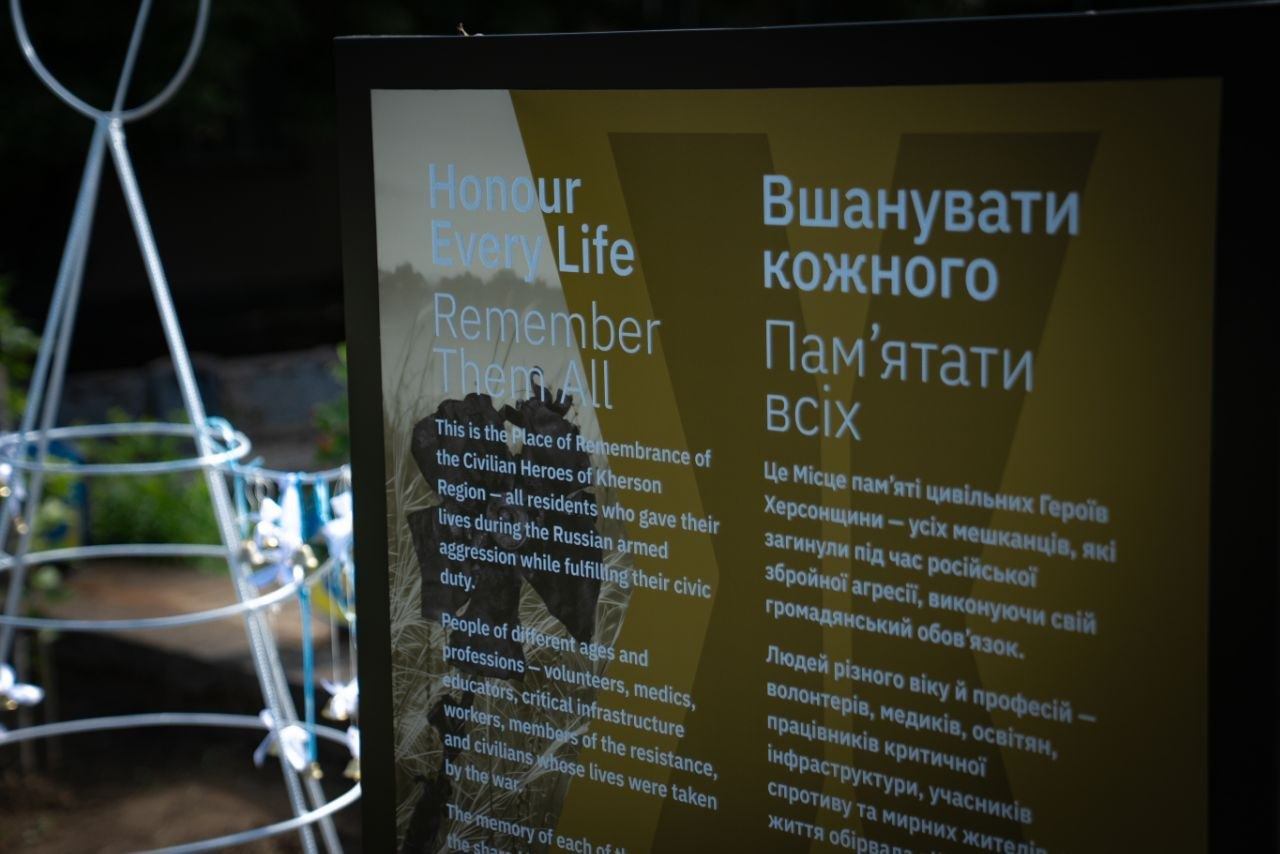 Інформаційний стенд меморіалу цивільним героям Херсонщини. На темній панелі великими літерами нанесено напис англійською та українською мовами: «Вшанувати кожного. Пам’ятати всіх». Нижче розміщено текст про те, що це місце пам'яті присвячене мешканцям регіону різних професій та віку, які загинули під час російської агресії.