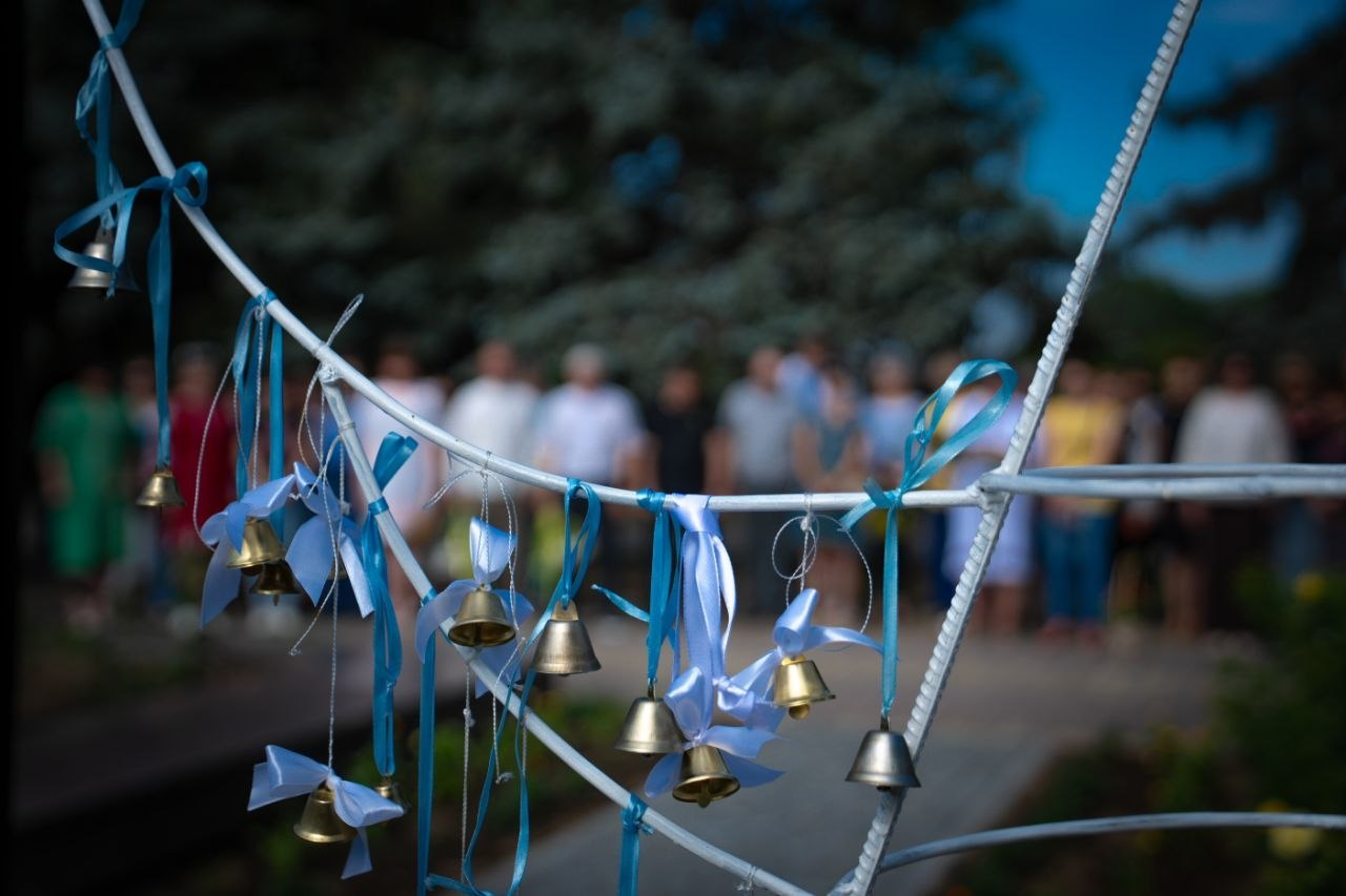 Маленькі дзвоники пам'яті на меморіальному заході. На білій металевій конструкції на передньому плані підвішено металеві дзвоники, прикрашені блакитними та синіми стрічками. На розмитому задньому плані видно шеренгу людей (учасників заходу та керівництво), які стоять просто неба на тлі дерев.