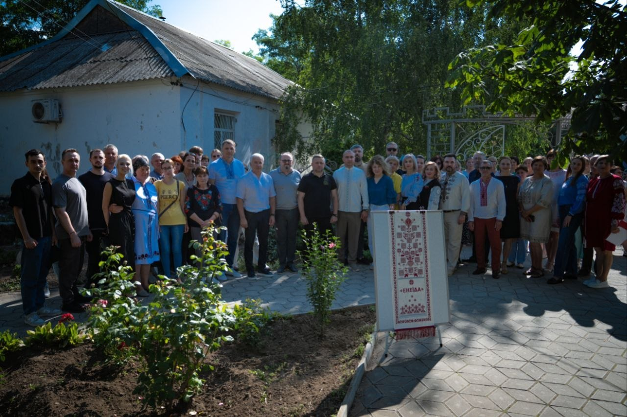 Учасники відкриття Місця пам’яті цивільних Героїв Херсонщини в селі Чорнобаївка. Велика група людей, серед яких представники обласної та місцевої влади, військовослужбовці та мешканці громади, вишикувалися для спільного фото на подвір’ї. На передньому плані праворуч встановлено інформаційний стенд із зображенням традиційного українського вишитого рушника та написом «Енеїда».