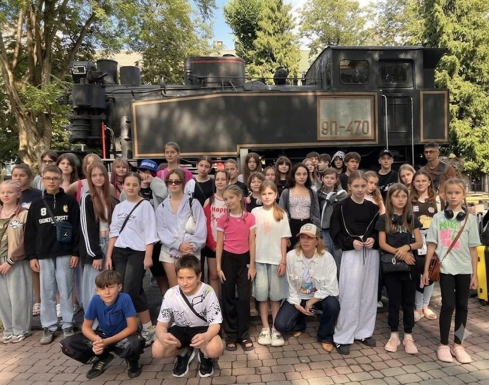 Група дітей та підлітків на екскурсії біля старовинного потяга. Велика група хлопців та дівчат у літньому одязі позує для спільного фото на бруківці. Двоє хлопців присіли на передньому плані, а позаду них стоять інші учасники разом із дорослою супроводжуючою в кепці. На задньому плані височіє великий чорний екскурсійний паровоз із бортовим номером «9П-470» в оточенні зелених дерев.