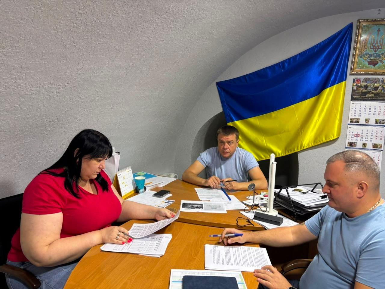 Робоча зустріч за участю Антона Самойленка в укритті. Заступник начальника Херсонської ОВА (по центру в блакитній футболці) разом із колегами вивчає документи за дерев’яним столом. Кабінет має закруглену білу стелю, а на стіні позаду розвішано великий прапор України та календар.