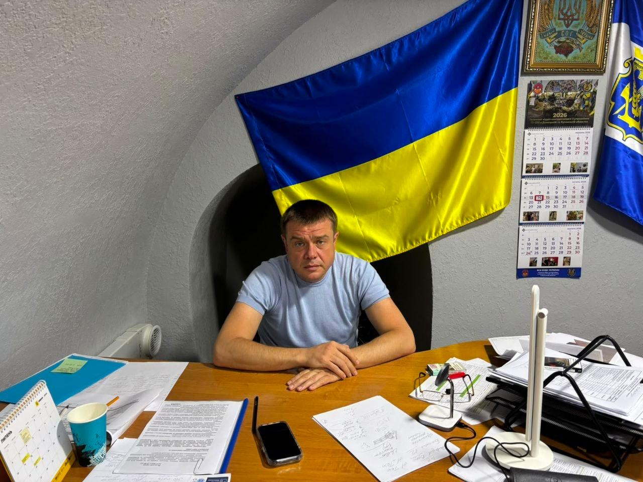 Антон Самойленко у робочому кабінеті-укритті. Заступник начальника Херсонської ОВА в блакитній футболці сидить за дерев'яним столом, на якому розкладені документи та папери. Позаду нього на стіні висить великий державний прапор України та календар.