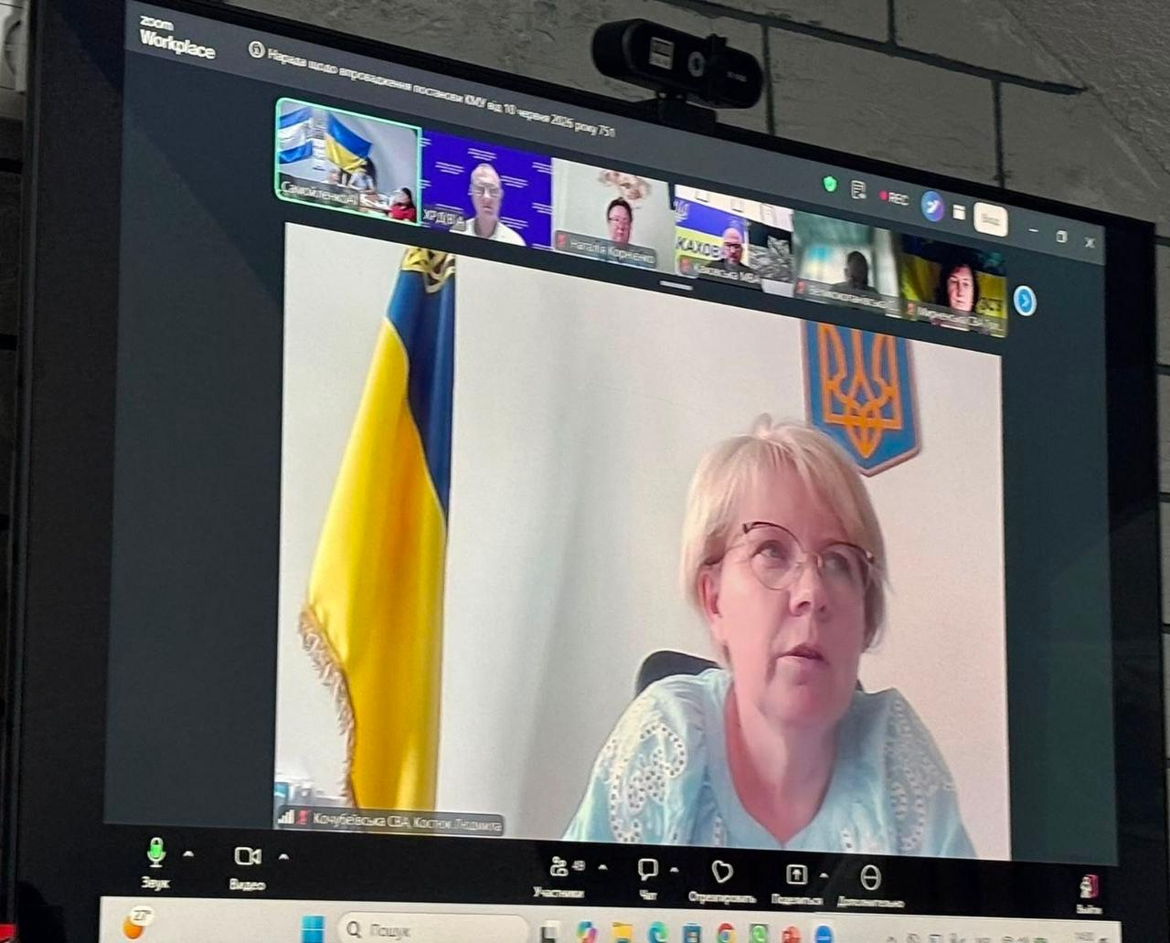 Онлайн-нарада у Zoom на великому екрані. Головне вікно відеозв'язку транслює виступ начальниці Кочубеївської СВА Людмили Костюк. Жінка в окулярах та світлій блузці говорить на тлі державного прапора та герба України. У верхній частині екрана видно віконця інших учасників наради, серед яких Каховська МВА, заступник начальника Херсонської ОВА Антон Самойленко та інші посадовці.