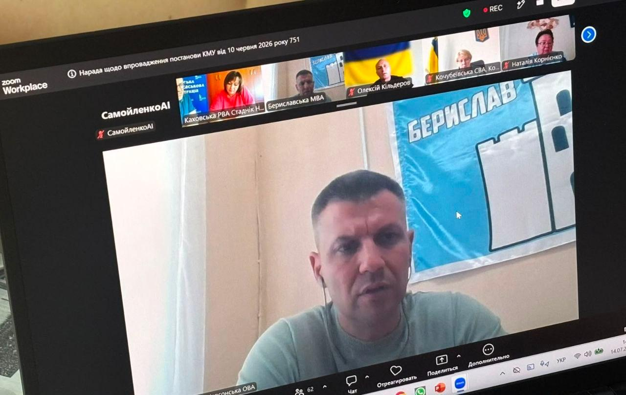 Олександр Алчієв виступає на онлайн-нараді у Zoom. Екран ноутбука транслює виступ начальника Бериславської МВА на тлі блакитного прапора з написом «Берислав». У верхньому рядку віконець видно інших учасників, зокрема Антона Самойленка. Зверху вказано тему зустрічі щодо впровадження постанови КМУ № 751.