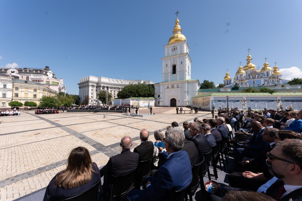 Офіційні урочистості на Михайлівській площі в Києві. Знімок зроблено зі спини гостей заходу, які сидять у глядацьких рядах просто неба. По центру площі облаштовано зону для виступів. На задньому плані велично підноситься Михайлівський Золотоверхий собор зі своєю дзвіницею, а ліворуч видно будівлю Міністерства закордонних справ України під яскравим синім небом.