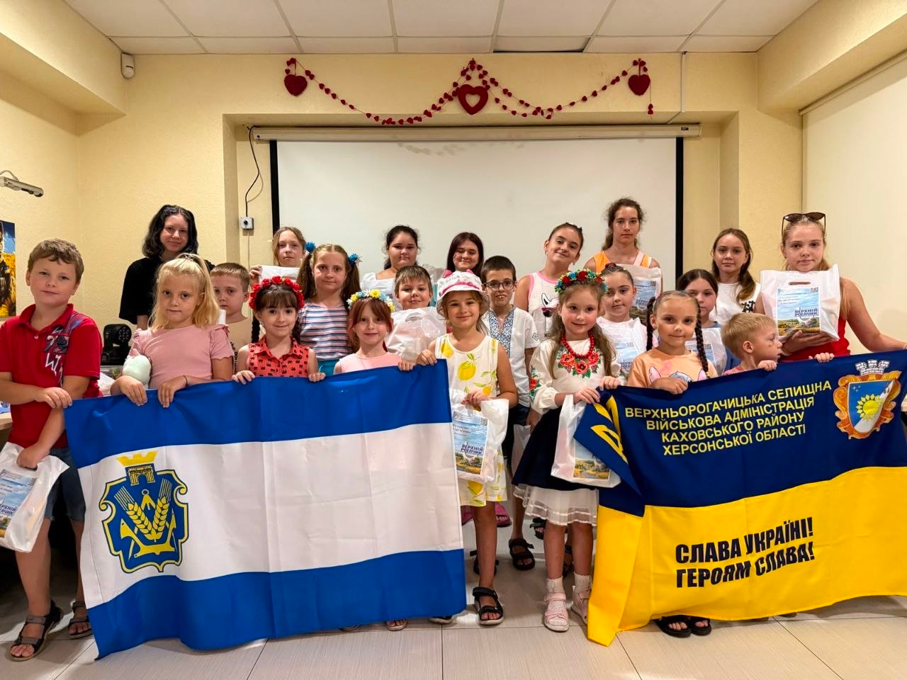 Велика група дітей та підлітків позує в приміщенні, тримаючи в руках пакети з подарунками. Попереду вони розгорнули два прапори: ліворуч — синьо-біло-синій прапор Херсонської області з її гербом, праворуч — синьо-жовтий стяг із написом «Верхньорогачицька селищна військова адміністрація Каховського району Херсонської області» та гаслом «Слава Україні! Героям Слава!».