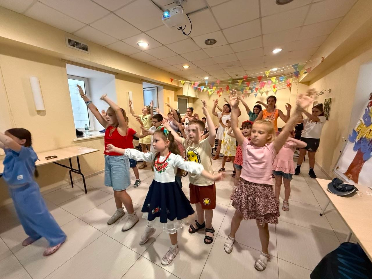 Велика група дітей та підлітків повторює рухи, піднявши руки вгору під час заходу. На передньому плані видно дівчинку в українському національному костюмі (вишиванці та спідниці) та інших дітей у літньому одязі. Стела кімнати прикрашена різнокольоровими прапорцями-гірляндами.