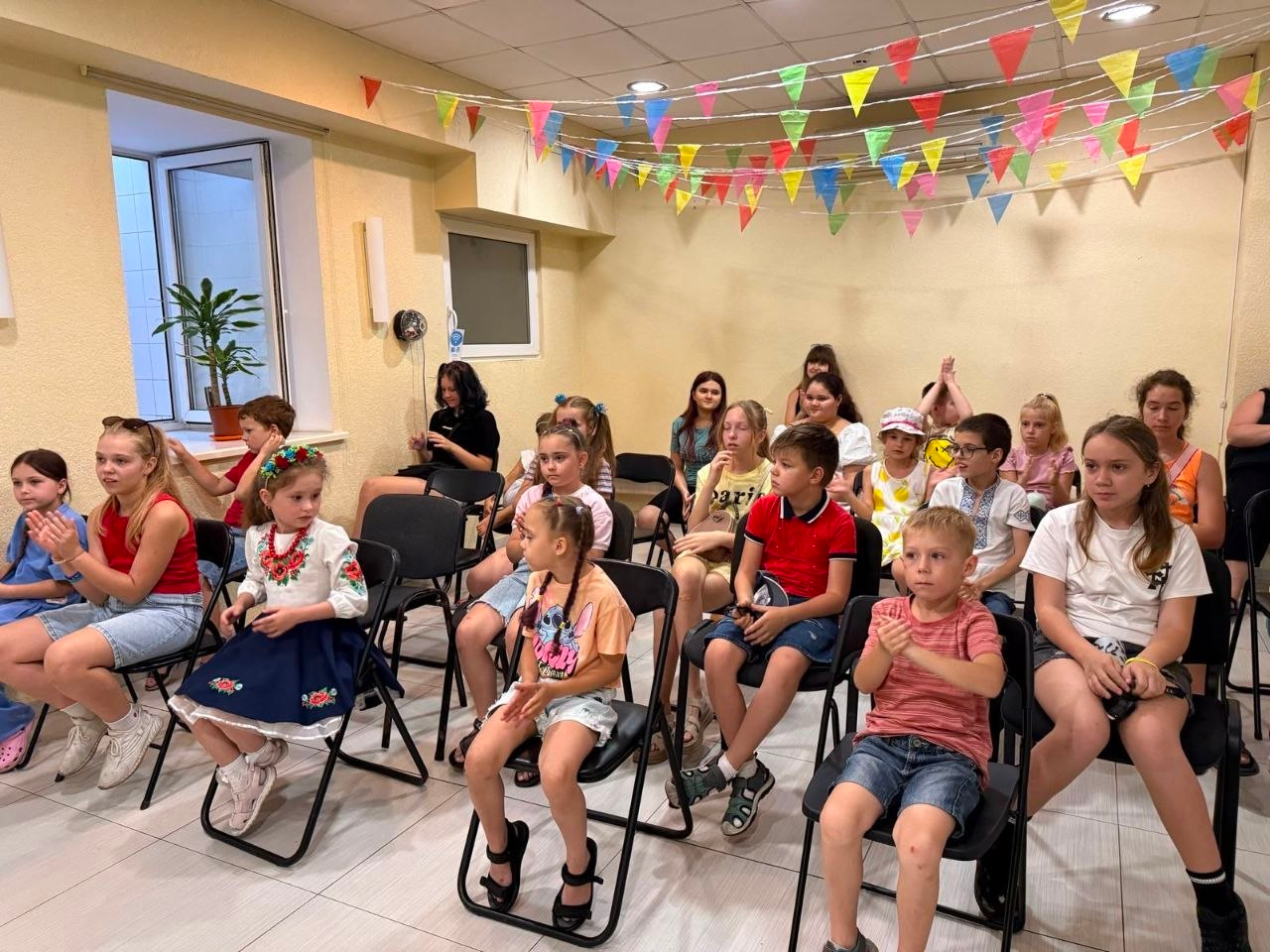 Група дітей та підлітків сидить на стільцях під час святкової програми в залі, прикрашеній кольоровими прапорцями. Ліворуч на передньому плані видно дівчинку в українській вишиванці та віночку, яка аплодує разом з іншими учасниками.