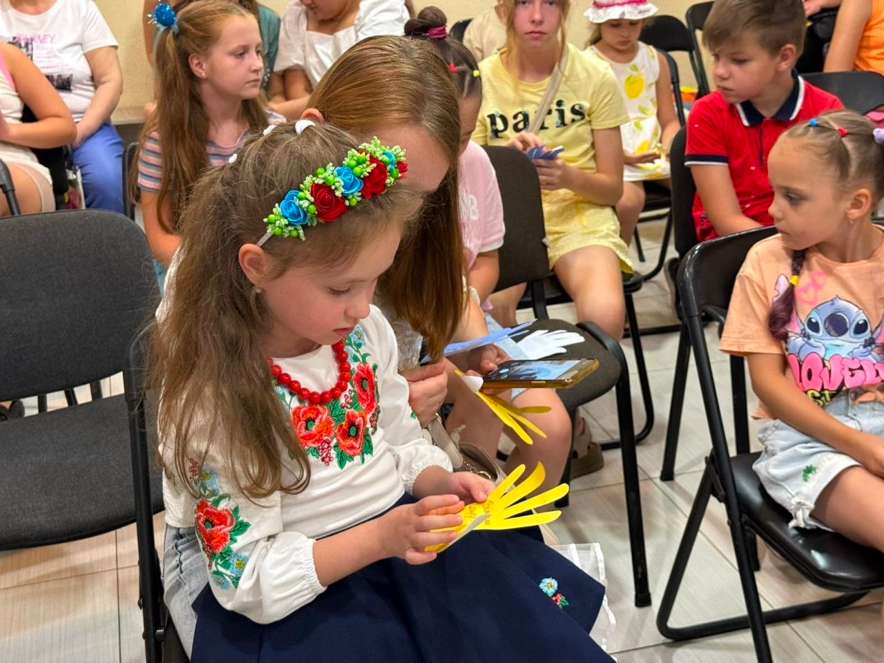На передньому плані засідання чи заходу сидить маленька дівчинка у вишитій сорочці та віночку, яка роздивляється вирізану з жовтого паперу долоньку з написом. Поруч і позаду неї на стільцях сидять інші діти-учасники патріотично-розважального заходу, які тримають сині та жовті паперові заготовки.