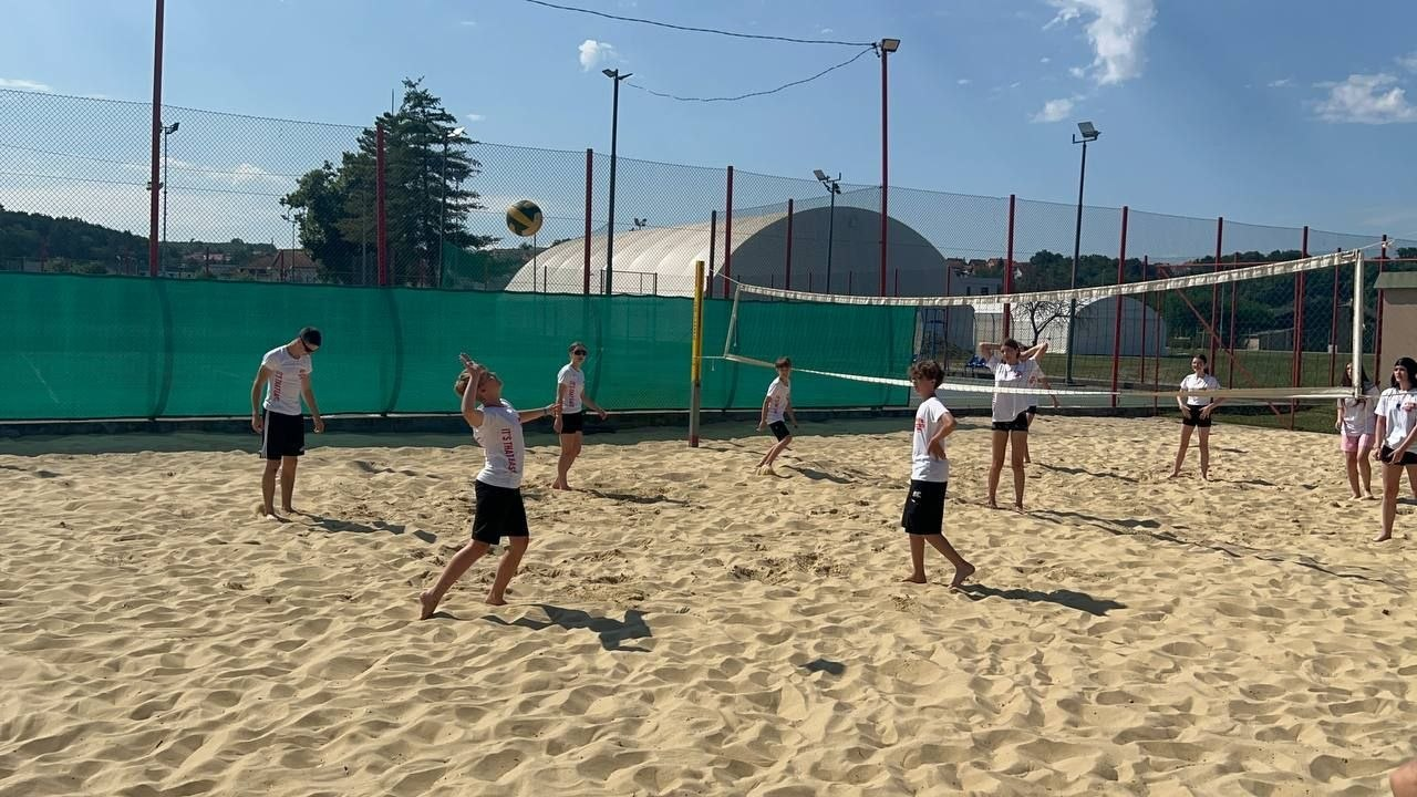 Група хлопців та дівчат у білих футболках розігрує волейбольний м'яч через сітку просто неба. На передньому плані зліва підліток замахнувся, щоб відбити м'яч, який завис у повітрі. Інші учасники гри стежать за подачею, перебуваючи на піску.