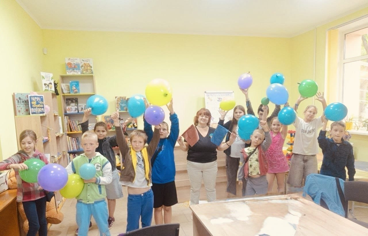 Група дітей разом із жінкою-організаторкою позують у світлій кімнаті з жовтими стінами. Учасники тримають у піднятих руках багато різнокольорових повітряних кульок (синіх, зелених, жовтих та фіолетових). Ліворуч на задньому плані видно книжкові полиці з дитячою літературою.