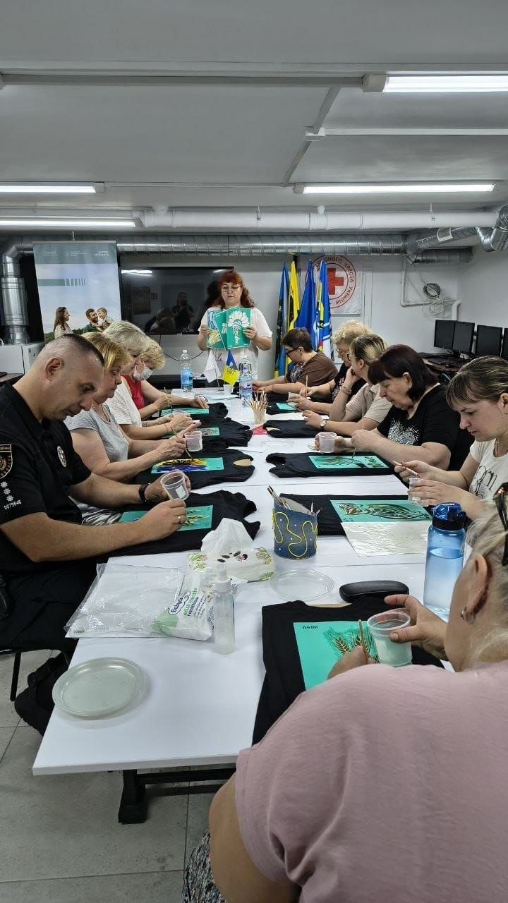 Група дорослих людей сидить за довгим столом у приміщенні та розмальовує чорні футболки під керівництвом жінки-інструктора, яка стоїть попереду.