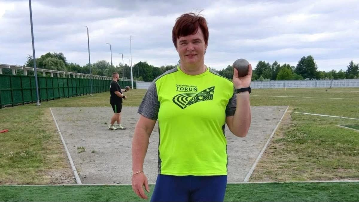 Українська легкоатлетка Тетяна Насонова у яскраво-салатовій футболці з написом «Torun» стоїть у секторі для метань на стадіоні, посміхається та тримає в лівій піднятій руці спортивне ядро.