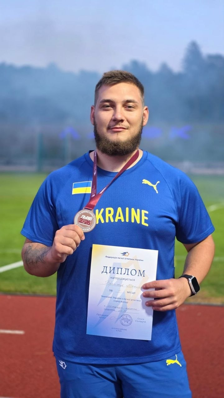 країнський легкоатлет Микита Брагінець у синій спортивній формі з жовтим написом «UKRAINE» стоїть на стадіоні, тримаючи в одній руці медаль на шиї, а в іншій — офіційний диплом Федерації легкої атлетики України за третє місце серед молоді в метанні молота вагою 7,260 кг.