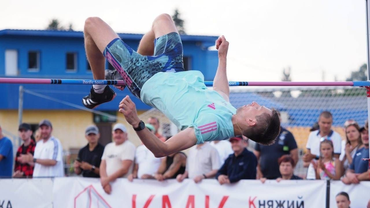 Український легкоатлет Андрій Проценко у світло-зеленій футболці виконує стрибок у висоту, перелітаючи спиною через планку на тлі глядачів та банера з написом «Княжий».