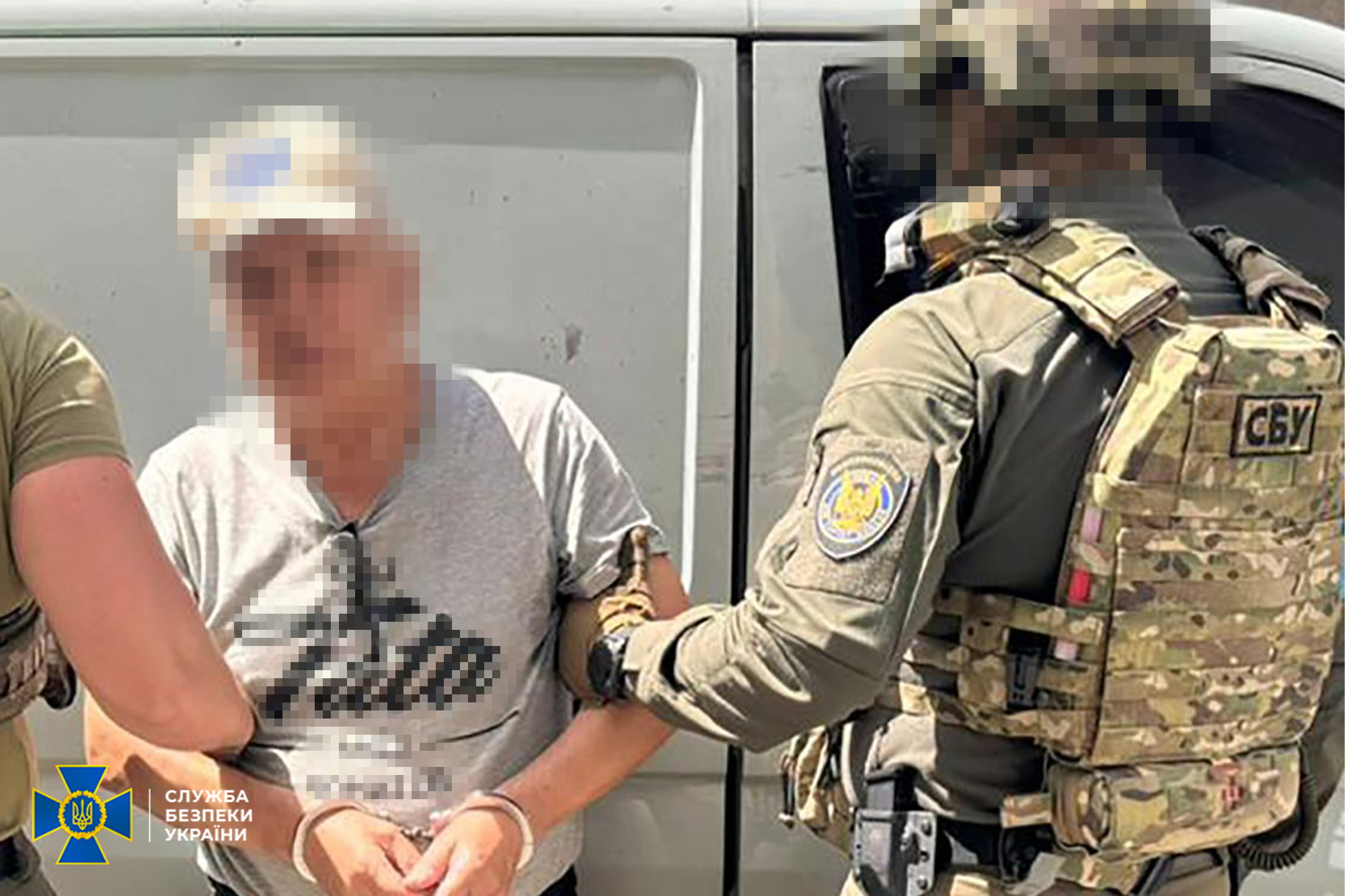 Співробітник Служби безпеки України у військовій формі та камуфляжному бронежилеті із шевроном «СБУ» затримує чоловіка в сірій футболці, застібаючи на його руках кайданки. Обличчя обох чоловіків розмиті за допомогою пікселів. У нижньому кутку фото розміщено офіційний логотип СБУ.