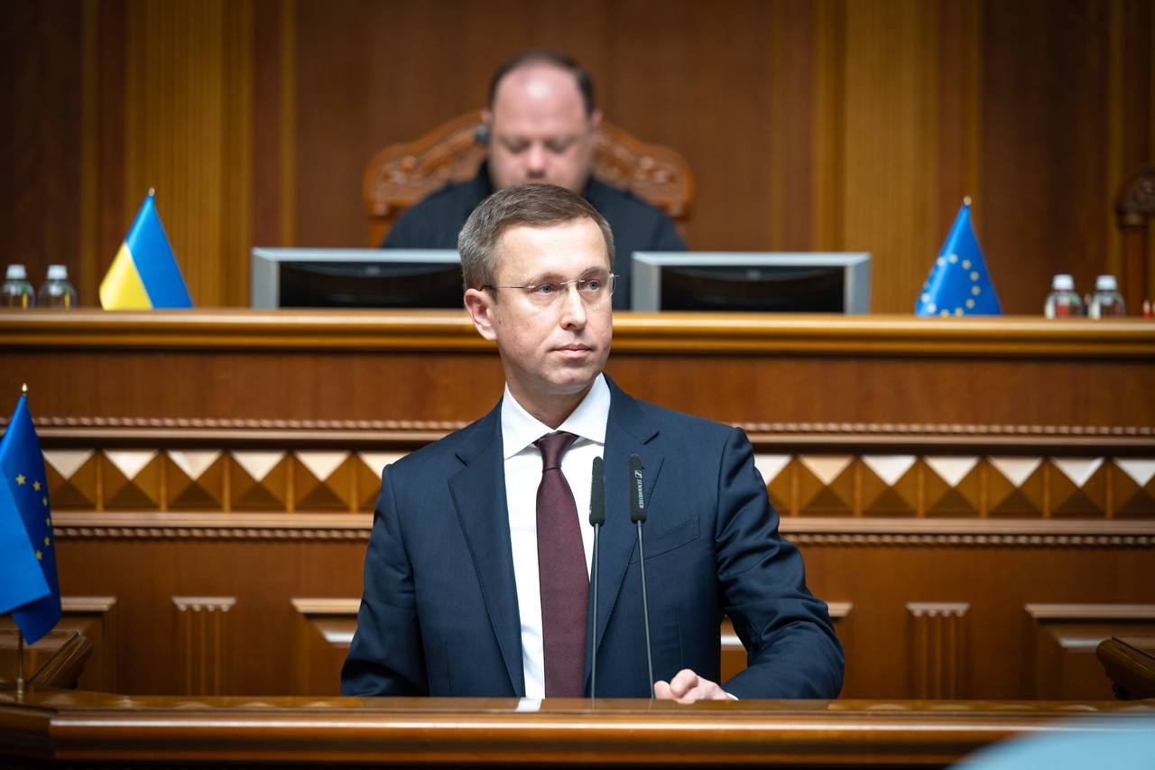 Прем’єр-міністр України Сергій Корецький у темно-синьому костюмі, білій сорочці та бордовій краватці виступає за трибуною у сесійній залі Верховної Ради України на тлі президії, державного прапора України та прапора Європейського Союзу.