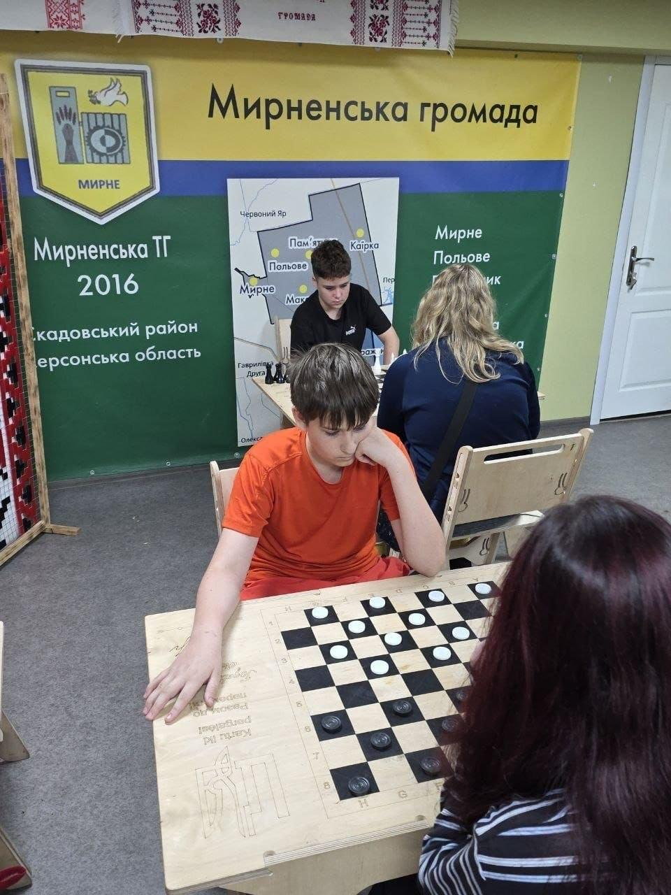 Хлопчик в помаранчевій футболці зосереджено грає в шашки за дерев'яним столом на тлі великого банера «Мирненська громада» Скадовського району Херсонської області, де на задньому плані інші учасники також грають у настільні ігри.
