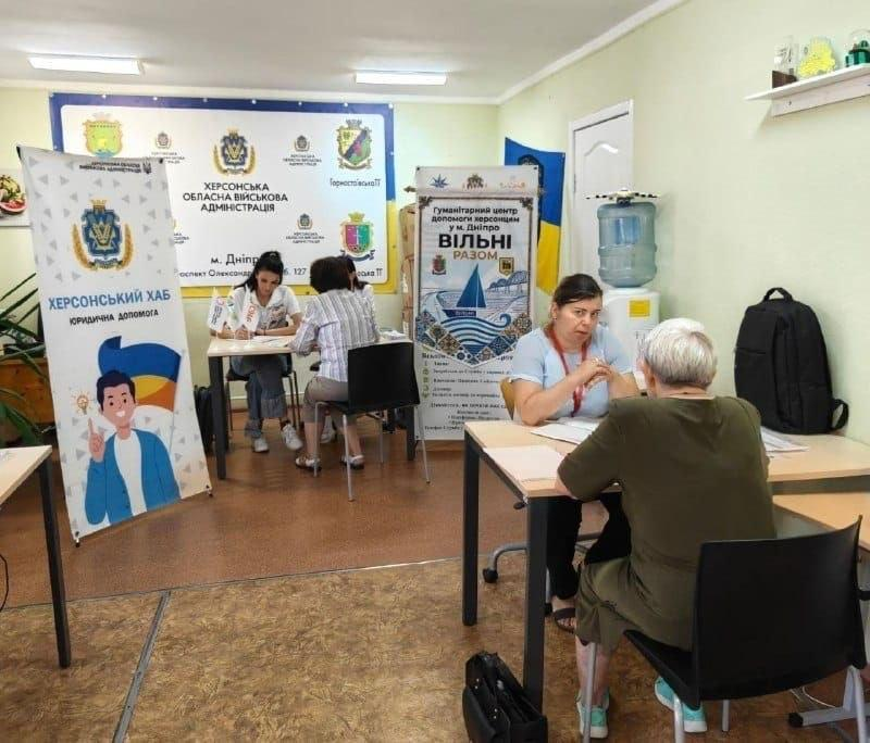 У приміщенні гуманітарного хабу «Вільні разом» у місті Дніпро працівники за столами консультують та надають юридичну допомогу переселенцям із Херсонської області на тлі банерів Херсонської ОВА.