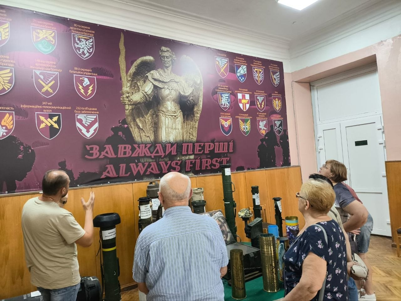 Переселенці з Херсонщини під час патріотичного заходу до Дня українських миротворців розглядають виставку тубусів від зброї та гільз у Житомирському Будинку офіцерів на тлі банера ДШВ «ЗАВЖДИ ПЕРШІ» із зображенням Архистратига Михаїла.